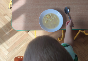 Na stole stoi talerz z zupą. Dziecko obok talerza kładzie łyżkę.