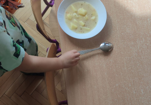 Na stole stoją talerze z zupą. Dziecko obok talerza kładzie łyżkę.