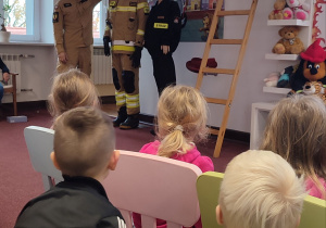 Dzieci siedzą na krzesełkach i słuchają strażaka, który opowiada o wyposażeniu strażaka.