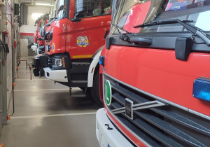 Wnętrze remizy strażackiej. Widok na kilka stojących obok siebie wozów strażackich.