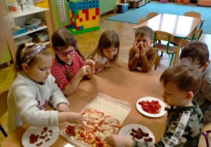 Dzieci siedząc przy stoliku układają ulubione dodatki na pizzy.
