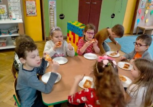 Dzieci siedząc przy stoliku jedzą upieczoną już pizzę.