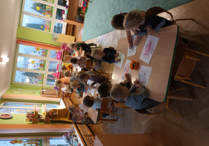 Dzieci siedzą przy stoliczkach i kolorują obrazek misia