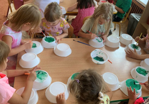Dzieci siedząc przy stolikach malują na zielono talerzyki