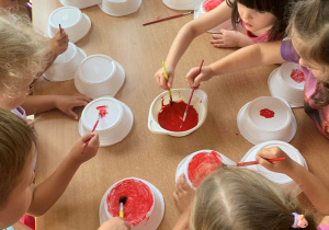 Dzieci siedząc przy stolikach malują na czerwono talerzyki