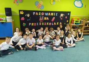 Grupowe zdjęcie dzieci na tle dekoracji z napisem "Pasowanie na przedszkolaka".