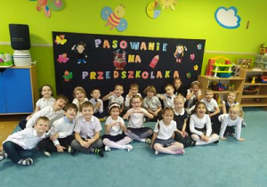 Grupowe zdjęcie dzieci na tle dekoracji z napisem "Pasowanie na przedszkolaka".