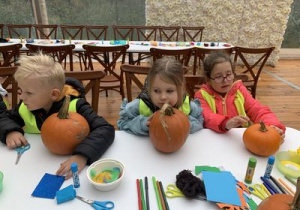 Dzieci siedzą przy stoliku z dyniami i elementami do ich ozdobienia.