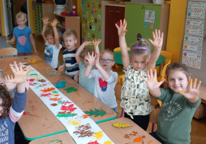 Dzieci podnoszą ręce do góry w tle widać ich pracę plastyczną