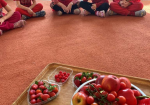 Dzieci siedzą w kole na dywanie - przed nimi ustawione są czerwone owoce i warzywa