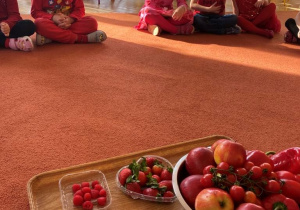 Dzieci siedzą w kole na dywanie - przed nimi ustawione są czerwone owoce i warzywa