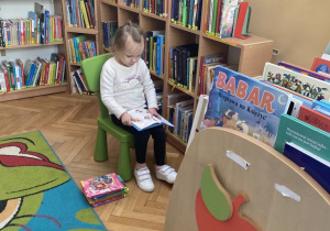 Dziewczynka siedząc na krzesełku wśród regałów bibliotecznych ogląda książeczkę