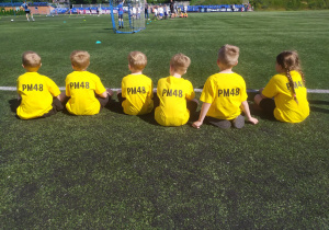 Dzieci siedzą tyłem, oczekując na mecz w koszulkach z nazwą przedszkola