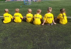 Dzieci siedzą na murawie boiska w koszulkach z logo przedszkola.