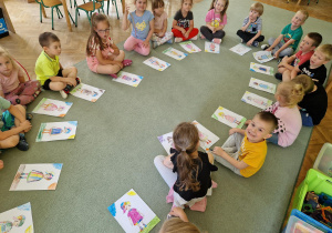 Dzieci siedzą w kole na dywanie i prezentują pokolorowane obrazki.