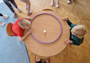 dzieci w parach na  przeciwko siebie wprowadzają w ruch piłeczkę na zmianę w obrębie hula hop.