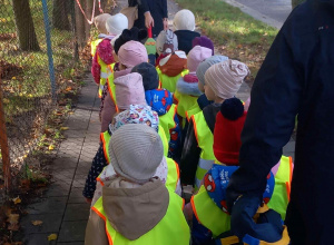 Dzieci ubrane w kamizelki odblaskowe wspólnie ze swoimi paniami idą na pobliskie skrzyżowanie 