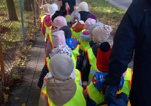 Dzieci ubrane w kamizelki odblaskowe wspólnie ze swoimi paniami idą na pobliskie skrzyżowanie