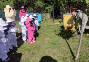 Dzieci ubrane w kombinezony stoją obok uli, słuchają co mówi pszczelarz i patrzą na "wejście" do ula