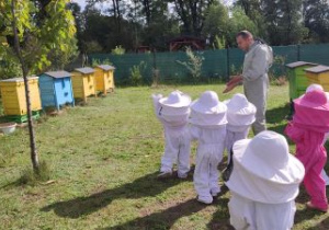 Dzieci ubrane w kombinezony stoją pośród uli i obserwują latające pszczoły