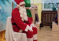 Mikołaj siedzi na krześle. Obok stoi Pani Elf. Przy nogach Mikołaja, na podłodze leży worek z prezentami.