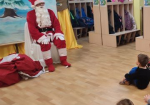 Przed dziećmi siedzi na krześle Mikołaj. Obok niego, na podłodze leży worek z prezentami