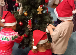  Dzieci ubrane w czerwone czapki zakładają bombki na świąteczne drzewko