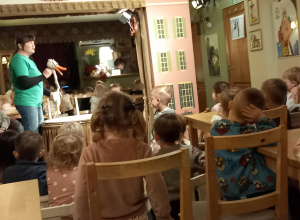 Dzieci siedzą na krzesłach i patrzą się w kierunku sceny, na której jest aktorka trzymająca kaczątko