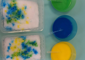 Na stole leżą pojemniki z zabarwionym śniegiem oraz kolorową wodą z pipetami