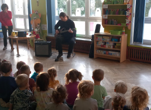 Dzieci siedzą na dywanie i słuchają jak pan gra na gitarze elektrycznej