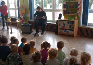 Dzieci siedzą na dywanie i słuchają jak pan gra na gitarze elektrycznej