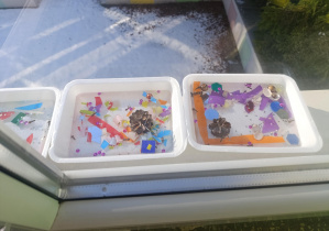 Tacki z wodą i brokatem, pomponikami, kolorowym papierem, szyszkami, muszelkami, cekinami stoją na zewnętrznym parapecie okna.