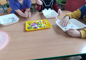 Dzieci układają kolorowy papier, cekiny, pomponiki na tackach.