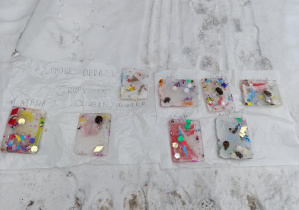 Zamarznięta woda wyjęta z tacek, a w niej umieszczone ozdoby: brokat, pomponiki, kolorowy papier, cekiny, szyszki.