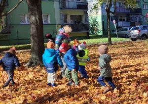 Dzieci biegają wśród drzew na trawniku zaspanym brązowymi, suchymi liśćmi.