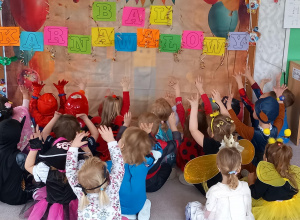 Dzieci siedzą w siadzie skrzyżnym i wskazują rękoma na napis bal karnawałowy
