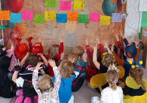 Dzieci siedzą w siadzie skrzyżnym i wskazują rękoma na napis bal karnawałowy