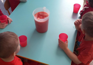 Dzieci siedzą przy stolikach z czerwonymi kubkami a na stole stoi dzbanek z sokiem pomidorowym