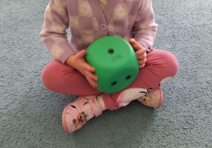 Dziewczynka siedzi w siadzie skrzyżnym i trzyma w rękach dużą piankową kostkę