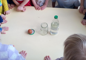 Dzieci stoją przed stołem, na którym stoi słoik, butelka z octem i jajko