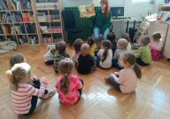 dzieci siedzą w bibliotece i  słuchają opowiadania czytanego przez panią bibliotekarkę