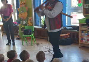 Dzieci siedzą na dywanie i patrzą jak pan gra na instrumencie