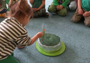 Dziewczynka wkłada w sztuczny tort zieloną świeczkę
