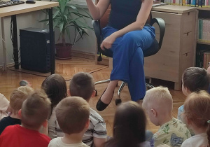 Pani bibliotekarka siedzi na krześle przed dziećmi i czyta im książkę.
