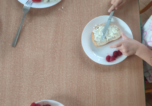 Na stole stoją talerze z kanapkami. Dzieci smarują kanapki białym serkiem.