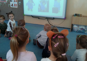 Dzieci siedzą na dywanie i oglądają prezentację na dużym ekranie