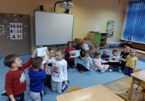 Dzieci siedzą na dywanie w grupach i pokazują ułożone obrazki misiów